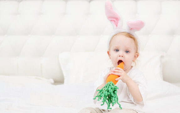 Little girl sitting on the bed in the bedroom with bunny ears on her head and a carrot in her hands