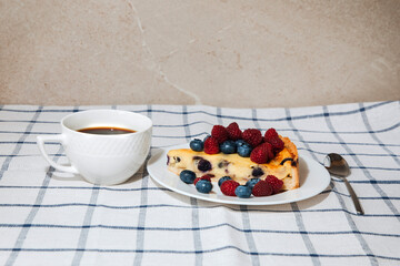 A piece of blueberry cheesecake decorated with raspberries and blueberries on the kitchen table with a cup of coffee. Front view