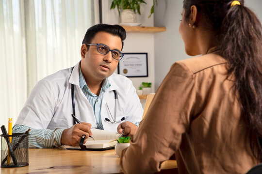 An Asian Indian male physician or man doctor sitting in a modern clinic wearing a stethoscope and apron writing drug prescription for a young female patient. Medical, medicine and healthcare concept