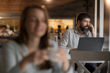 young businessman working on a laptop in cafe making a call on mobile phone, blurred girl sits in the foreground. Selective
