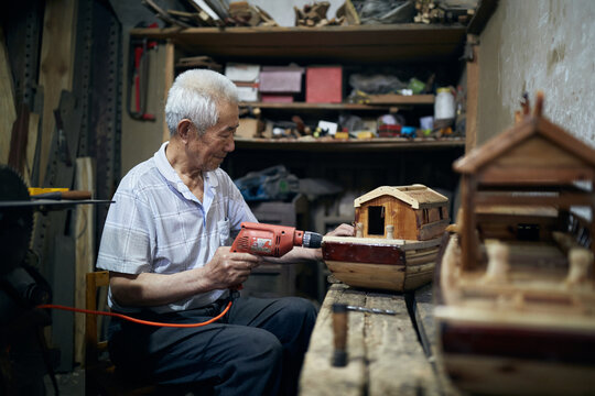 Older Asian Man Works On His Model Boats At His Workshop.