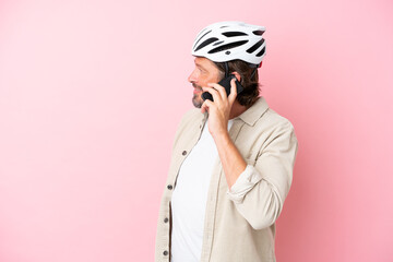 Senior dutch man with bike helmet isolated on pink background keeping a conversation with the mobile phone with someone
