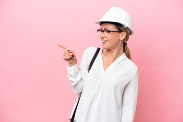 Young architect Russian woman with helmet and holding blueprints isolated on pink background pointing finger to the side and presenting a product