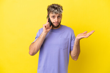 Young caucasian man using mobile phone isolated on yellow background having doubts while raising...