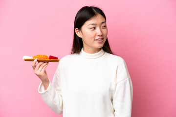 Young Chinese woman holding sashimi isolated on pink background looking to the side and smiling