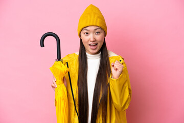 Young Chinese woman with rainproof coat and umbrella isolated on pink background celebrating a...
