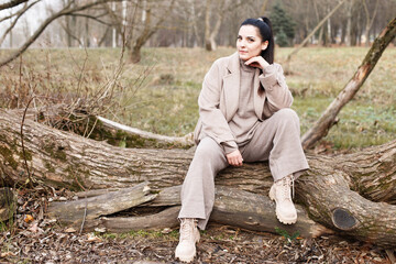Fototapeta premium portrait of brunette woman in beige coat with beautiful long hair sitting on a tree in the park