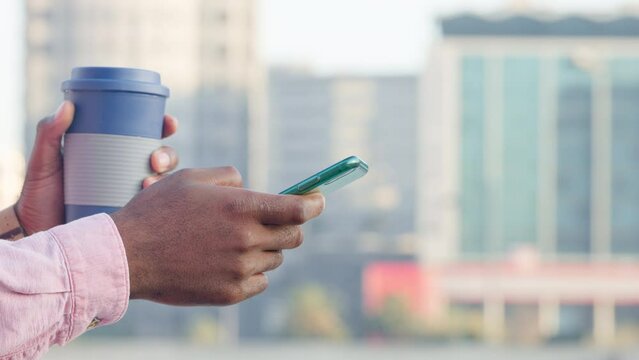Close up of a hand of an African American person scrolling on phone and holding cup with blurred background of buildings. Web developer texting client. IT professional taking break to drink coffee