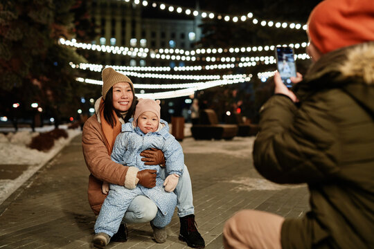 Horizontal shot of Asian man taking photos of his wife and toddler daughter on smartphone camera on winter evening