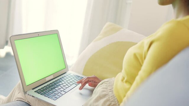 Woman scrolling and browsing online on a laptop with a green screen while relaxing on the couch at home. One person using social media or watching movies on the internet while sitting on the sofa