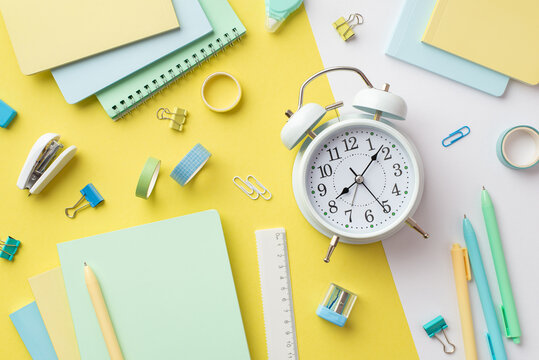 Back to school concept. Top view photo of alarm clock diaries sharpener ruler pens binder clips adhesive tape and stapler on bicolor yellow and white background
