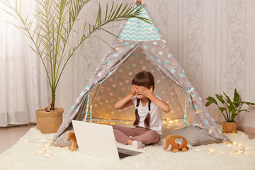 Indoor shot of little preschooler girl sitting in wigwam in front of notebook, having video call, covering eyes with palm, hiding herself, playing in new peetee tent. © sementsova321