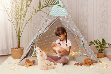 Indoor shot of little cute girl playing with soft toy teddy bear near the wigwam in the children's room, funny cute small female child having fun at home in peetee tent. © sementsova321
