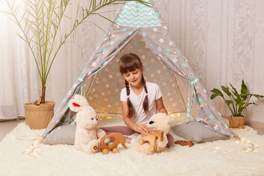 Indoor shot of charming dark haired Caucasian little girl wearing white t shirt sitting on floor in wigwam and playing with teddy bear, soft rabbit and eco wooden toys.