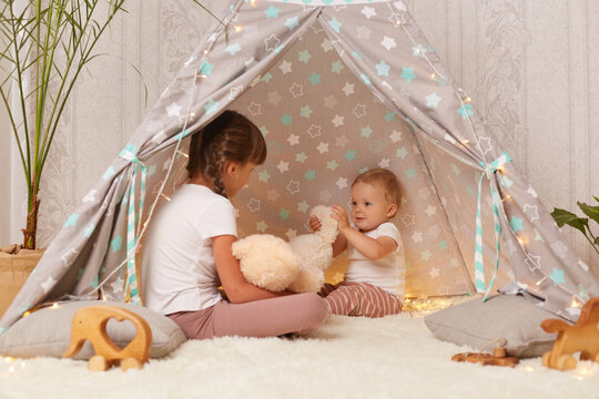 Image of little sisters playing in peetee tent with soft teddy bear, baby kid looking at toy with great interest, sibling sitting on floor on soft carpet, spending time together.