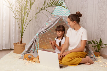 Portrait of dark haired female and her daughter wearing white t shirts sitting in wigwam in front of laptop, child using smart phone, mother looking at kid, family spending time together. © sementsova321