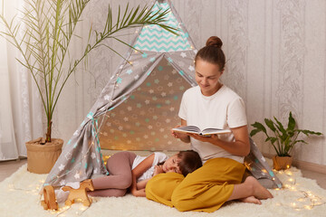 Portrait of beautiful young adult mother holding book and reading fairy tale for her little daughter, kid laying on floor and trying to fall asleep, family spending evening together.