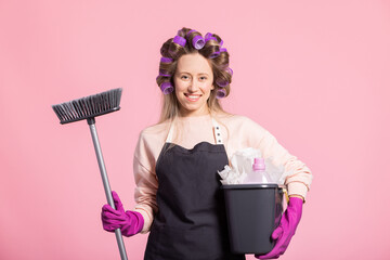 A young woman working from home arranges hair with rollers, wearing an apron, holding a broom for...