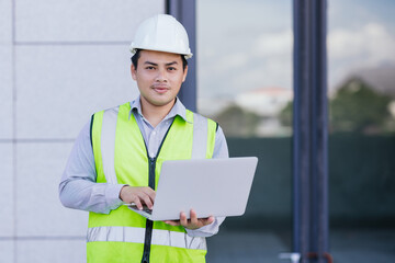 Asian engineer young man wearing safety vest and helmet standing using computer for check work on building construction site background. Engineering construction worker concept.