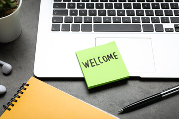 Paper note with word Welcome, laptop and notebook on grey table, closeup