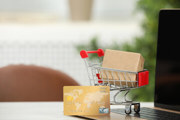 Online payment concept. Small shopping cart with bank card, box and laptop on white table