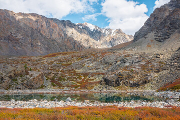 Turquoise mountain lake among autumn multicolor flora on rocks against high rocky mountain top in sunlight under clouds in blue sky. Sunlit alpine lake and large mountain range. Vivid autumn colors.
