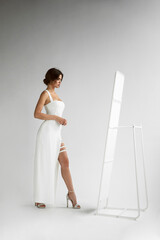Fototapeta na wymiar Full length view of young woman with hairstyle in white long dress posing sensual in studio, looking at mirror, isolated grey background.