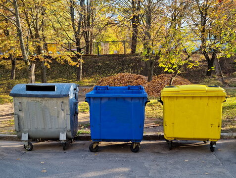 Yellow, blue and gray containers for separate garbage collection in the courtyard of an apartment building. Fallen leaves gathered in a pile on a sunny autumn day.