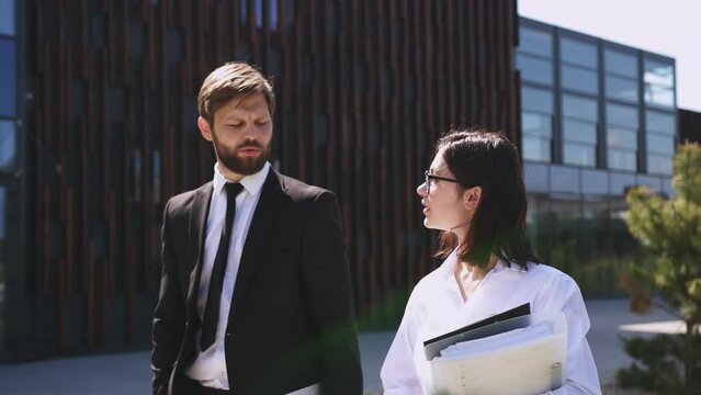 Handsome Caucasian male boss in suit and tie walking the street together with beautiful female assistant with documents and talking about work. Man employer and woman employee discussing job.