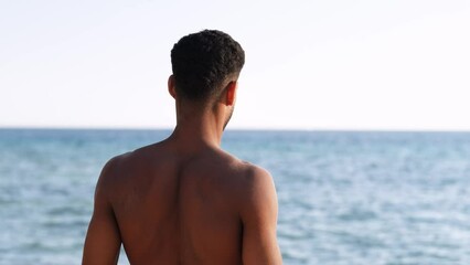 Backside view of relaxing young african man traveler on the beach, Handsome and confident topless guy looking on ocean sea, enjoying holidays.