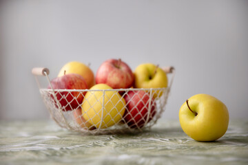 Pile of yellow and red apples lying on the table in a wire basket with white background and one apple lies nearby. Yellow and red apples in a basket on a table