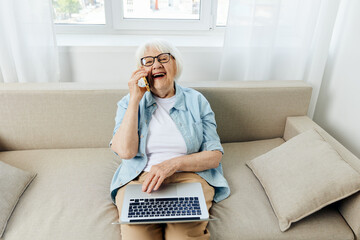 a joyful woman is sitting relaxed on the couch enjoying a conversation on the phone holding a laptop on her lap while working from home