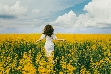 Fototapeta premium A girl in a white dress and curly hair runs across a field of yellow flowers.