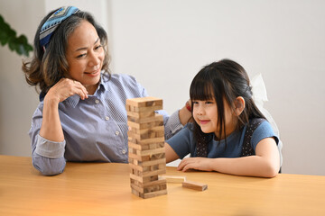 Cute little grandchild and grandmother having fun playing wood block stacking board game together in living room