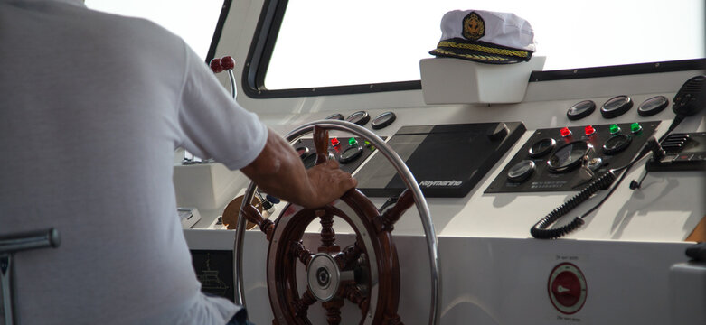 The captain of the ship holds the helm with his hand and the captain's cap lies nearby. White cockpit with brown steering wheel.