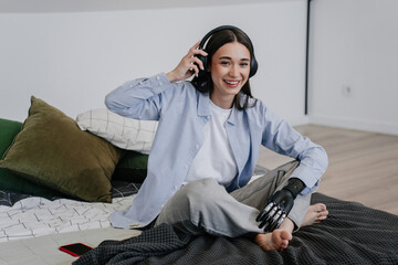 Brunette young woman with disability sitting on bed at home, with  bionic hand prosthesis, smiling,...