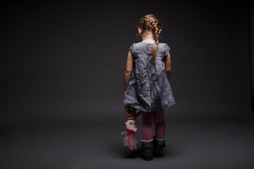 Rear image of a little sad girl with doll, toddler, offended by someone, being in bad mood, over dark grey background. - Powered by Adobe