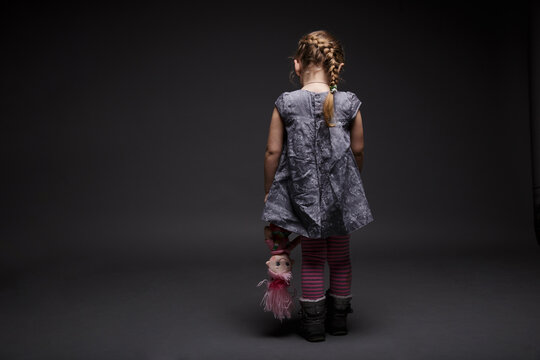 Rear image of a little sad girl with doll, toddler, offended by someone, being in bad mood, over dark grey background.