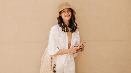 Fototapeta premium Smiling caucasian young woman looking at camera enjoying time outdoors during day. Brunette with smartphone in her hands wears casual clothes. Concept of use