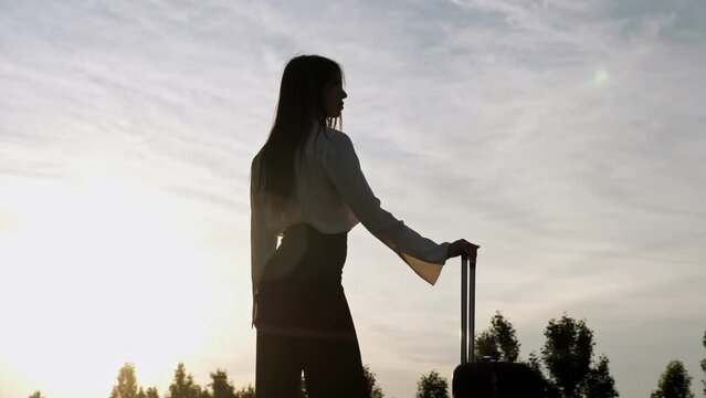 Silhouette of woman with long dark hair in business clothes stands holding suitcase at back sunset. Brunette looks at cloudy sky low angle shot