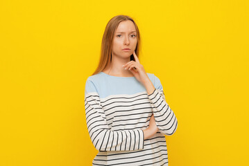 Obraz premium Portrait of thoughtful female, keeps finger on cheek, contemplates about future wedding with boyfriend or making important decision, dressed in casual striped sweatshirt, yellow background
