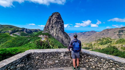 Sport man with backpack standing in front of massive volcanic rock formation Roque de Agando in Garajonay National Park on La Gomera, Canary Islands, Spain, Europe. Hiking trail on sunny day in summer
