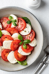 Caprese salad with sliced tomatoes, mozzarella, basil, olive oil with cutlery and salt shaker on white background close up. Top view