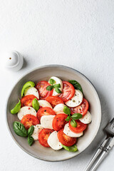 Italian caprese salad with sliced tomatoes, mozzarella, basil, olive oil with cutlery and salt shaker on white background with copy space. Top view