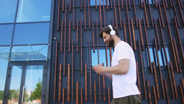Handsome young Caucasian man in sportswear and headphones walking the street, listening to music and texting mesage on smartphone. Male messaging online on mobile phone while stepping. Outdoors.