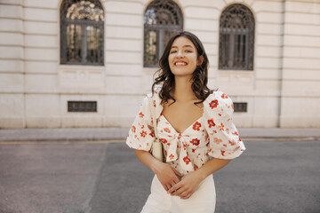 Happy young caucasian woman with snow-white smile looks at camera relaxing outdoors. Brunette wears white blouse and trousers in spring. Positive emotions concept
