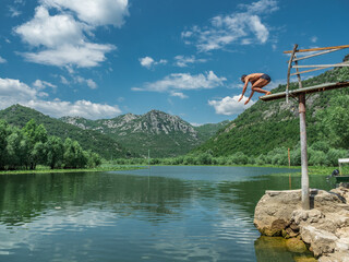 homem negro pulando em um lago em Montenegro. Com céu azul, nuvens e montanhas