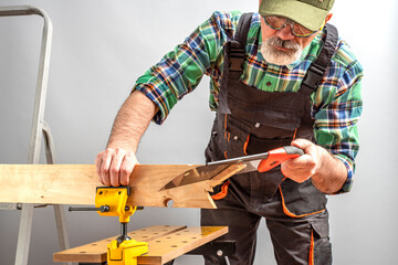 Aged worker working with a vise screwed to sawhorse. Senior man in overalls, cap and goggles