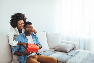 Shot of a loving husband giving his wife a gift. Boyfriend surprise his beautiful girlfriend with present while she is sitting on the sofa in the living room at home.