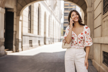 Positive young caucasian girl talking on phone spending time outdoors. Brunette wears white blouse, trousers in spring weather. Telephone technology concept